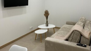 A modern living room with a flat-screen TV mounted on the wall, a beige sofa with a throw blanket, a small white table with a vase, and a white chair. - HOLLYWOOD RIVIERA (Cannes)