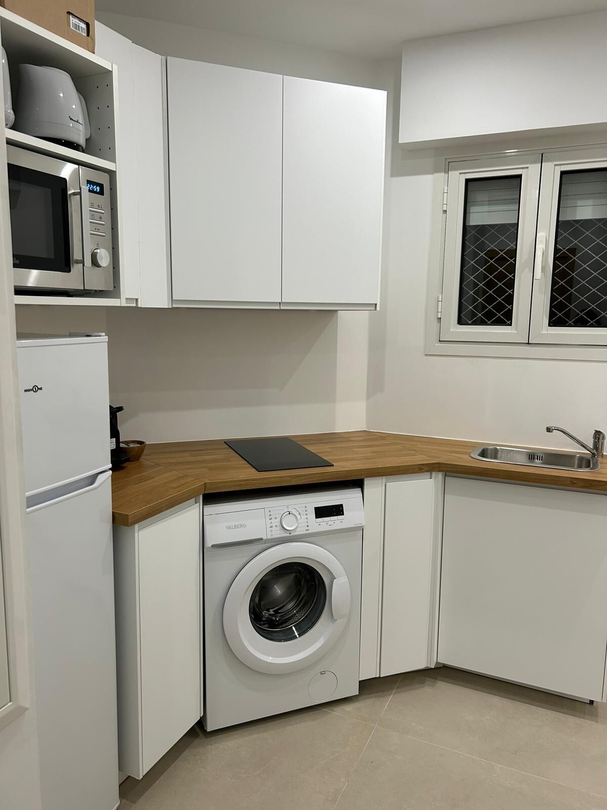 A compact kitchen with white cabinetry, a built-in microwave, a washing machine, and a window with a metal screen.