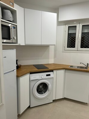 Une cuisine compacte avec des meubles blancs, un four à micro-ondes encastré, un lave-linge et une fenêtre dotée d’un moustiquaire métallique.