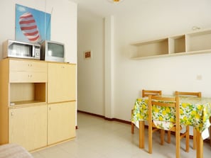 A room with a wooden cabinet containing a microwave and a television, a dining table with chairs, and a wall with a shelf above the table. - Cozy Beachside Apartment With Balcony (San Michele al Tagliamento)