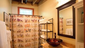 Un baño con techo de madera, un lavabo con pileta, inodoro, repisa con toallas y cortina de ducha con estampado de gallinas.