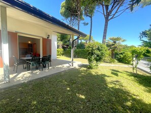 Eine Terrasse mit Tisch und Stühlen, ein überdachter Sitzbereich und Blick auf einen gepflegten Rasen und Bäume.