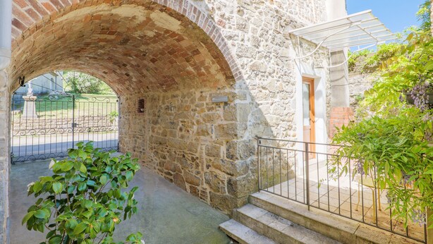 Un corredor de piedra con arco y escaleras que conducen a una puerta, rodeado de vegetación y un portón de hierro forjado.
