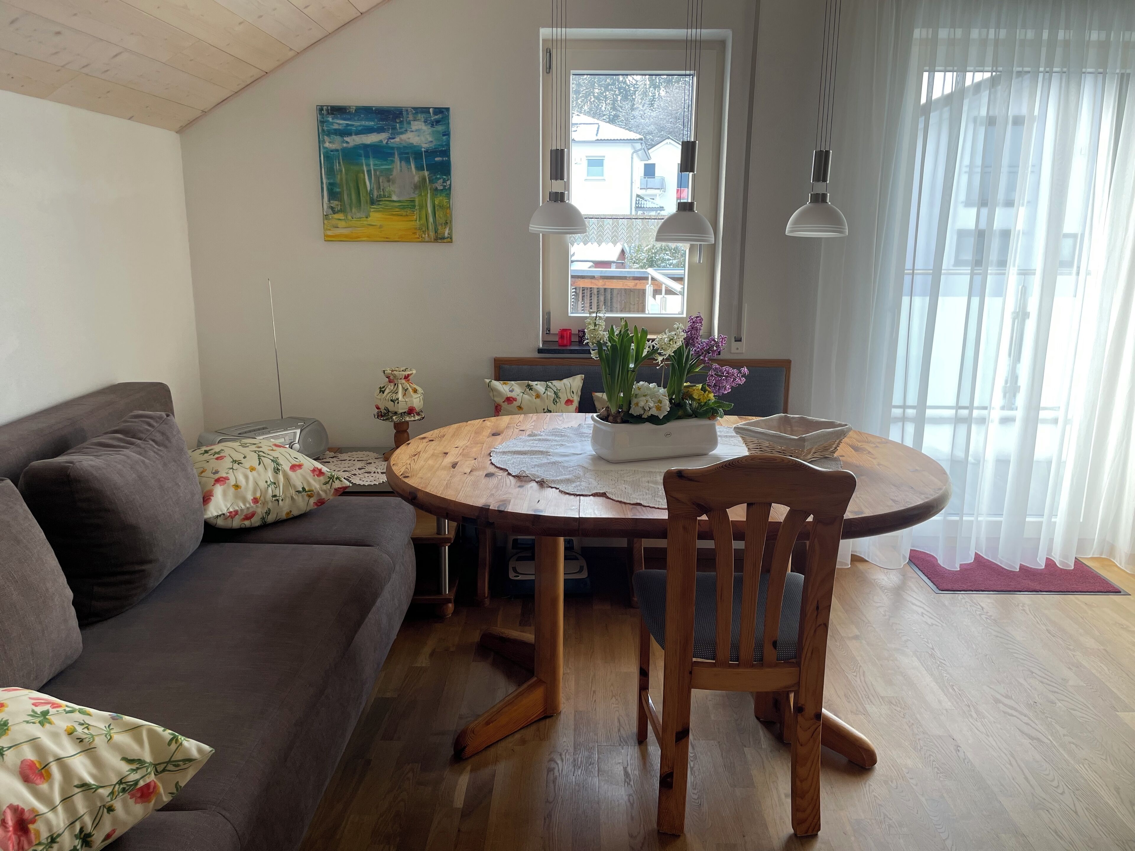 A cozy living room with a wooden dining table, a sofa, and a chair. There's a painting on the wall and a window with curtains.