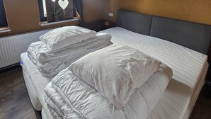 Un lit bien fait, avec du linge de lit blanc et des coussins décoratifs, une table de chevet avec une lampe, et une vue sur la verdure à travers une fenêtre ornée de découpes en forme de cœur.
