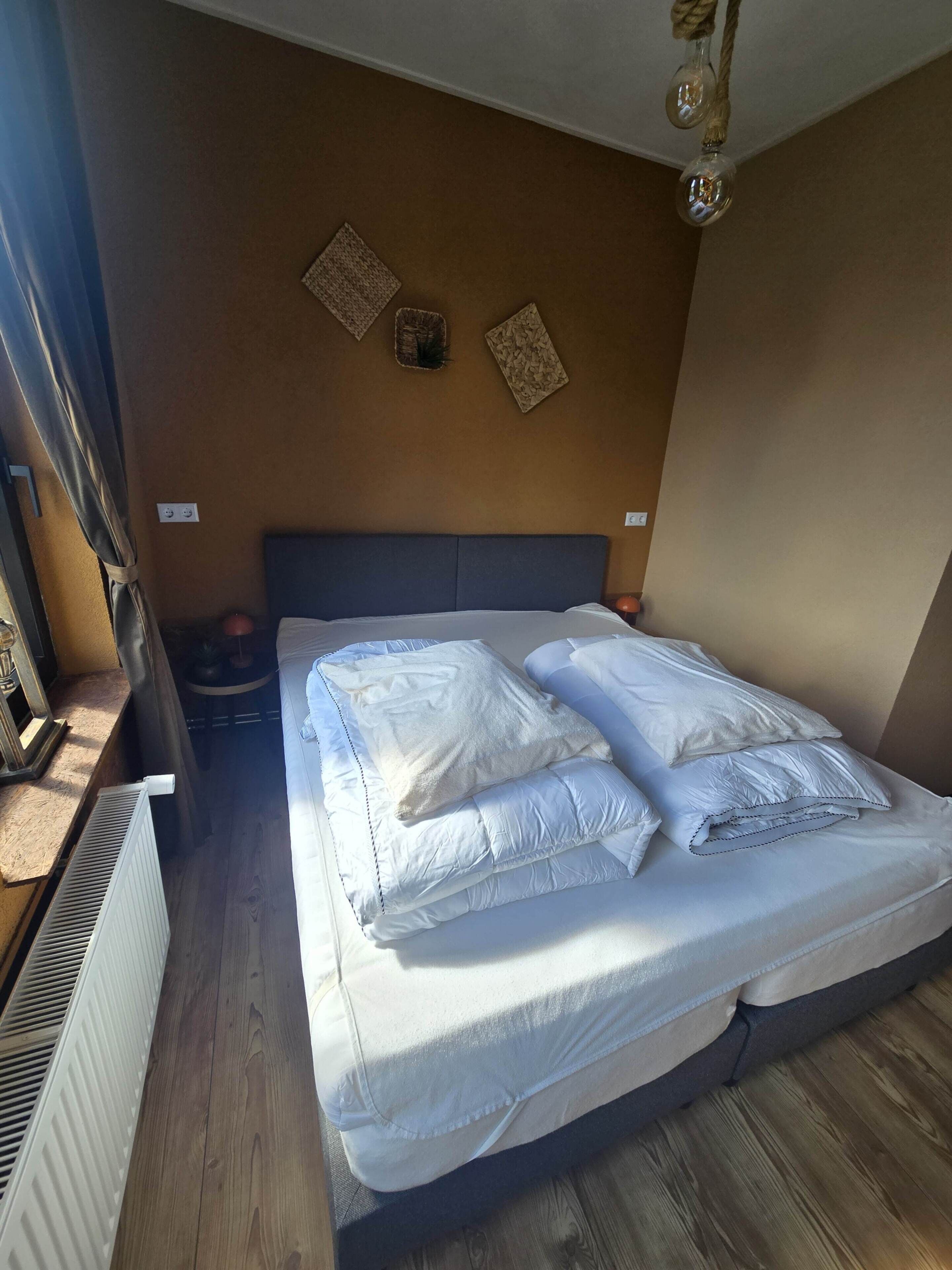 A neatly made bed with white linens and pillows, a wooden floor, a radiator, and a wall with two decorative items.