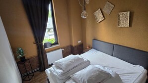Une chambre d’hôtel avec un lit, une fenêtre avec des rideaux, un radiateur, une plante en pot et des œuvres d’art accrochées au mur.
