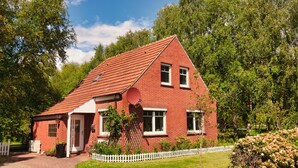 A red brick house with a white picket fence, a paved driveway, and a garden with various plants and flowers. - Idyllically situated apartment Logafeld in the Leeraner Meeden, pet friendly! (Leer (Ostfriesland))