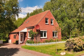 A red brick house with a white picket fence, a paved driveway, and a garden with various plants and flowers.