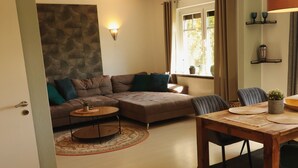 A living room with a sofa, a coffee table, a dining table with chairs, a stone accent wall, and a window with curtains. - Idyllically situated apartment Logafeld in the Leeraner Meeden, pet friendly! (Leer (Ostfriesland))