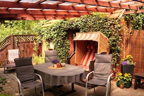 Eine Terrasse im Freien mit einem runden Tisch, vier Stühlen, einer Korusbänklage und einer mit Efeu bewachsenen Mauer.