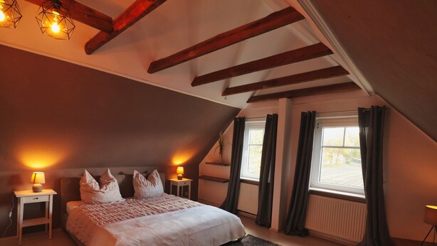 A bedroom with a sloped ceiling, a bed with pillows, bedside lamps, a window with curtains, and a radiator. - Idyllically situated apartment Logafeld in the Leeraner Meeden, pet friendly! (Leer (Ostfriesland))