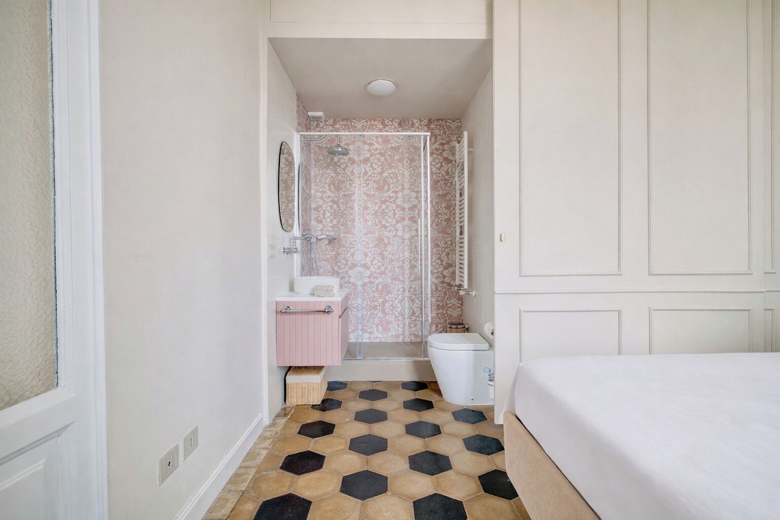 Un pasillo estrecho que conduce a un baño con ducha, inodoro y lavabo. El suelo presenta un diseño geométrico de baldosas.