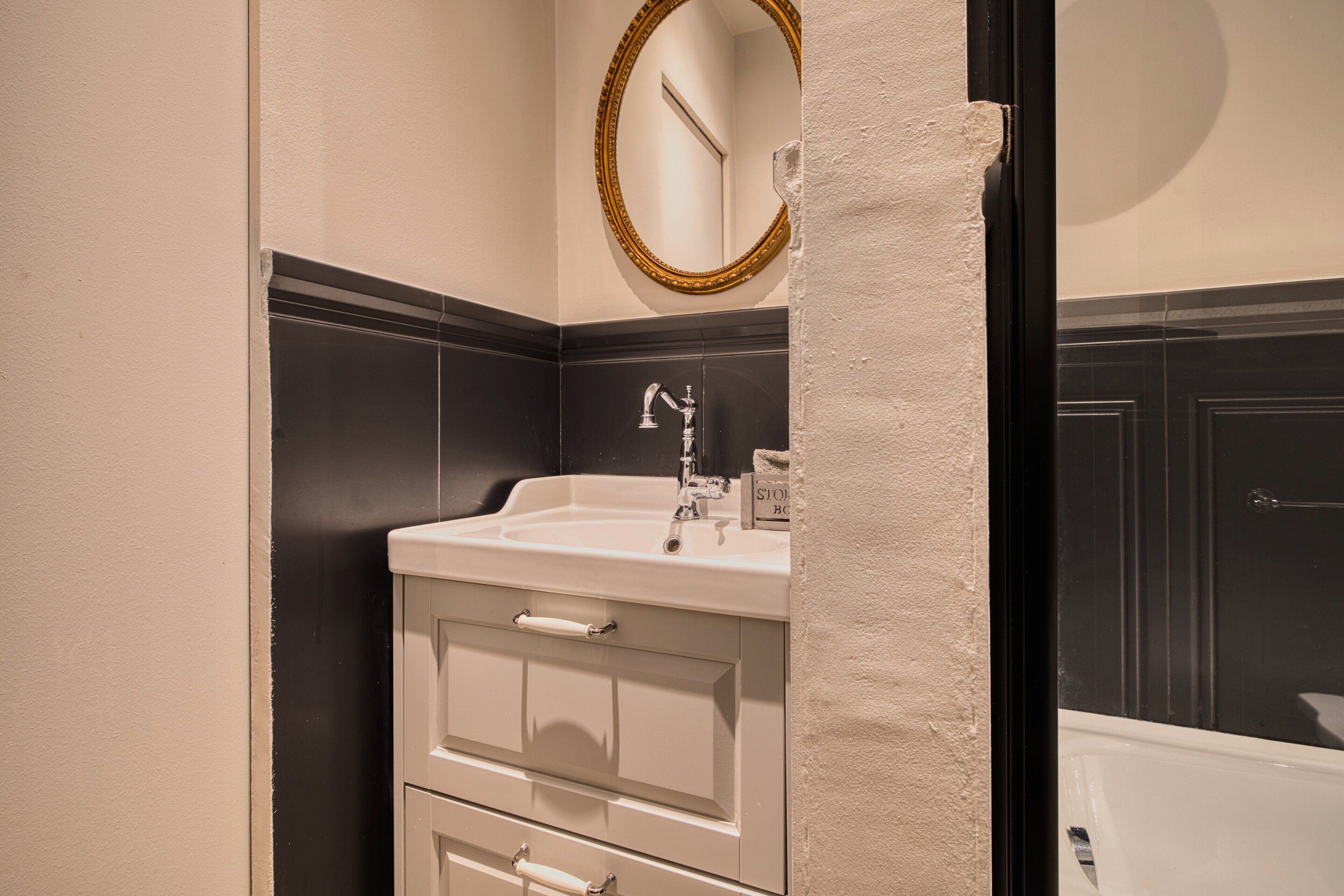 Un baño con un lavamanos blanco, paredes de azulejos blanco y negro, un espejo con marco dorado y una puerta negra.