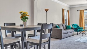 Ein modernes Wohnzimmer mit einem grauen Sofa, Türkis-Farbenen Sesseln, einem Esstisch mit Stühlen und einer Topfpflanze.