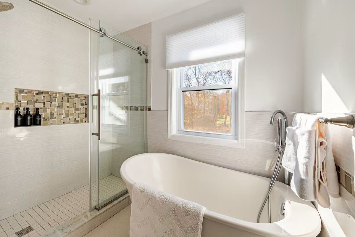 Un baño moderno con ducha de vidrio, bañera independiente y ventana con vista a árboles.
