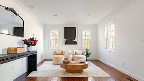Un salon spacieux doté d’un parquet, de deux fauteuils beiges, d’une table basse ronde et d’un grand miroir placé au-dessus d’une console sur laquelle se trouve un vase rempli de fleurs rouges.
