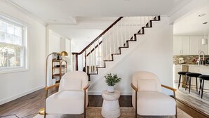 Un salon moderne avec deux fauteuils blancs, une petite table ronde agrémentée d’une plante, un escalier avec des marches en bois et un coin cuisine avec des tabourets de bar.