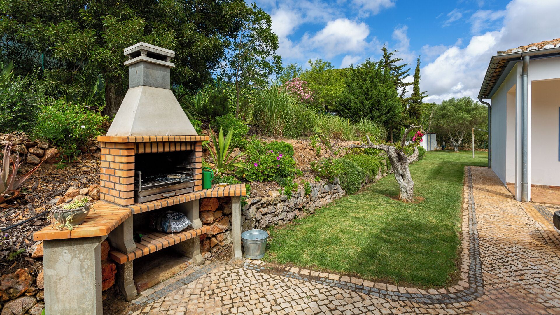 An outdoor brick-built barbecue area with a stone wall, a metal bucket, and a small garden with various plants and a tree.