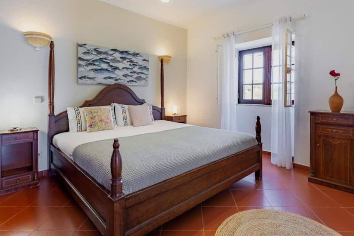A bedroom with a large bed, wooden headboard, two bedside tables, a nightstand, a vase with a flower, and a window with curtains.