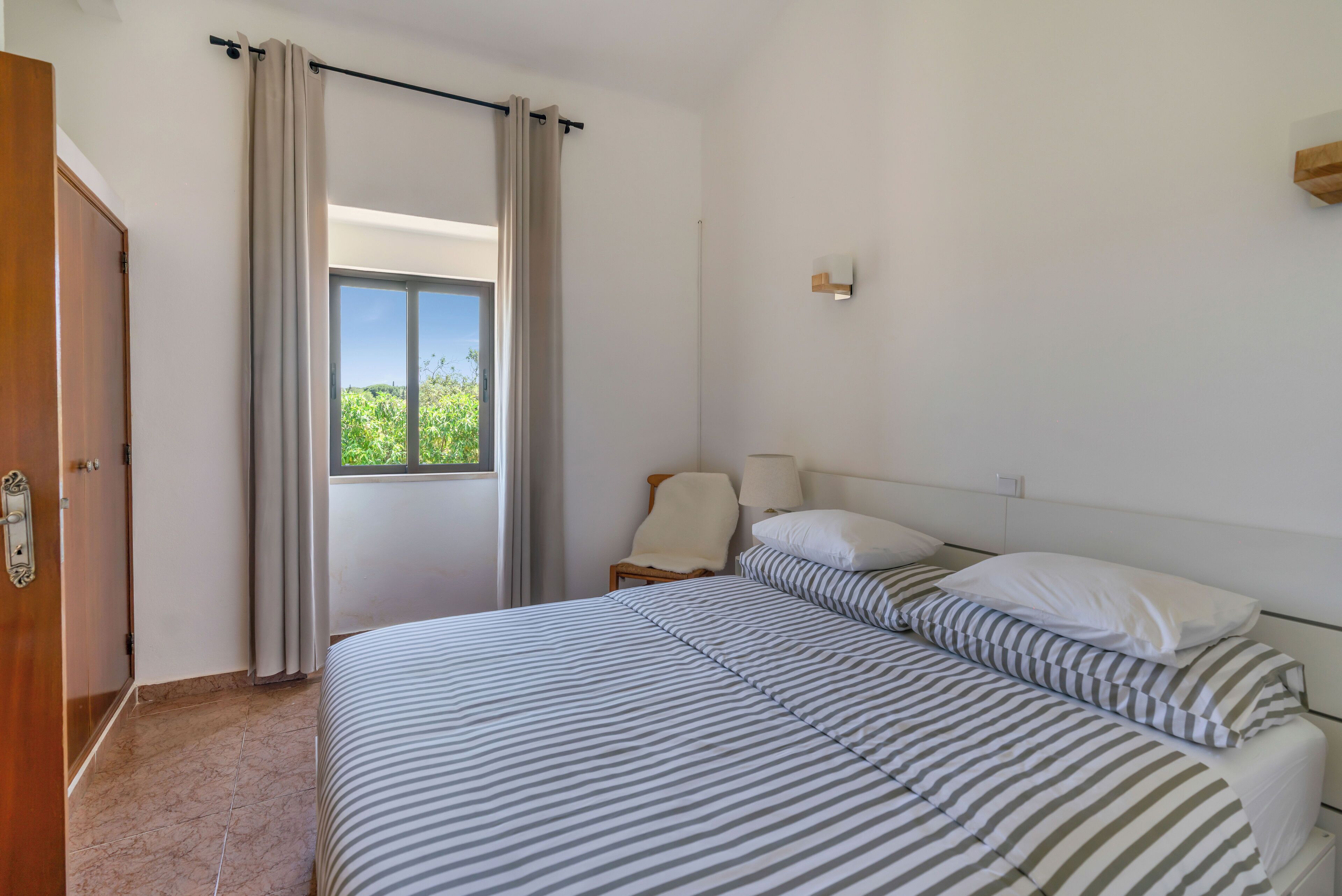 Une chambre d’hôtel avec un lit, une chaise, une fenêtre avec des rideaux et une lampe fixée au mur.
