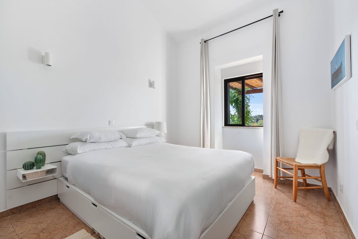 A modern bedroom with a large bed, a small bedside table, a chair, and a window with curtains.