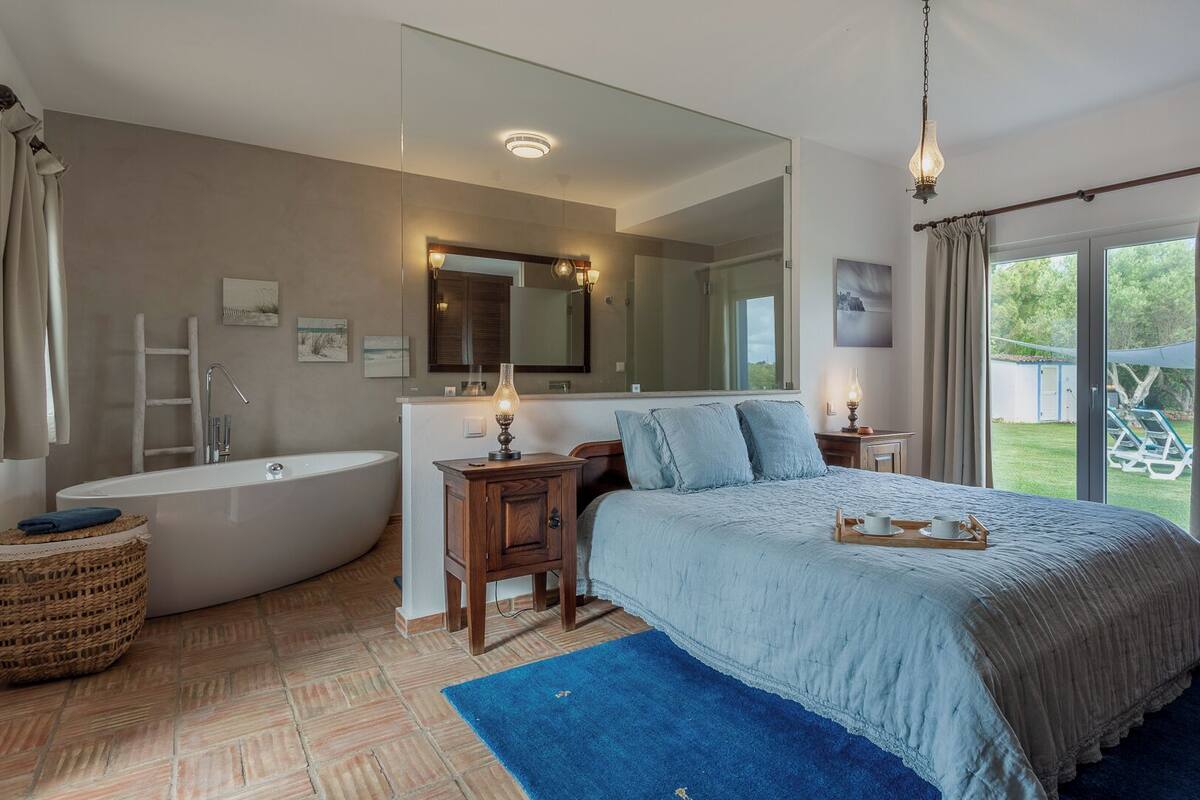 A bedroom with a large bed, a bathtub, a wooden cabinet, a mirror, and a view of the outdoors.