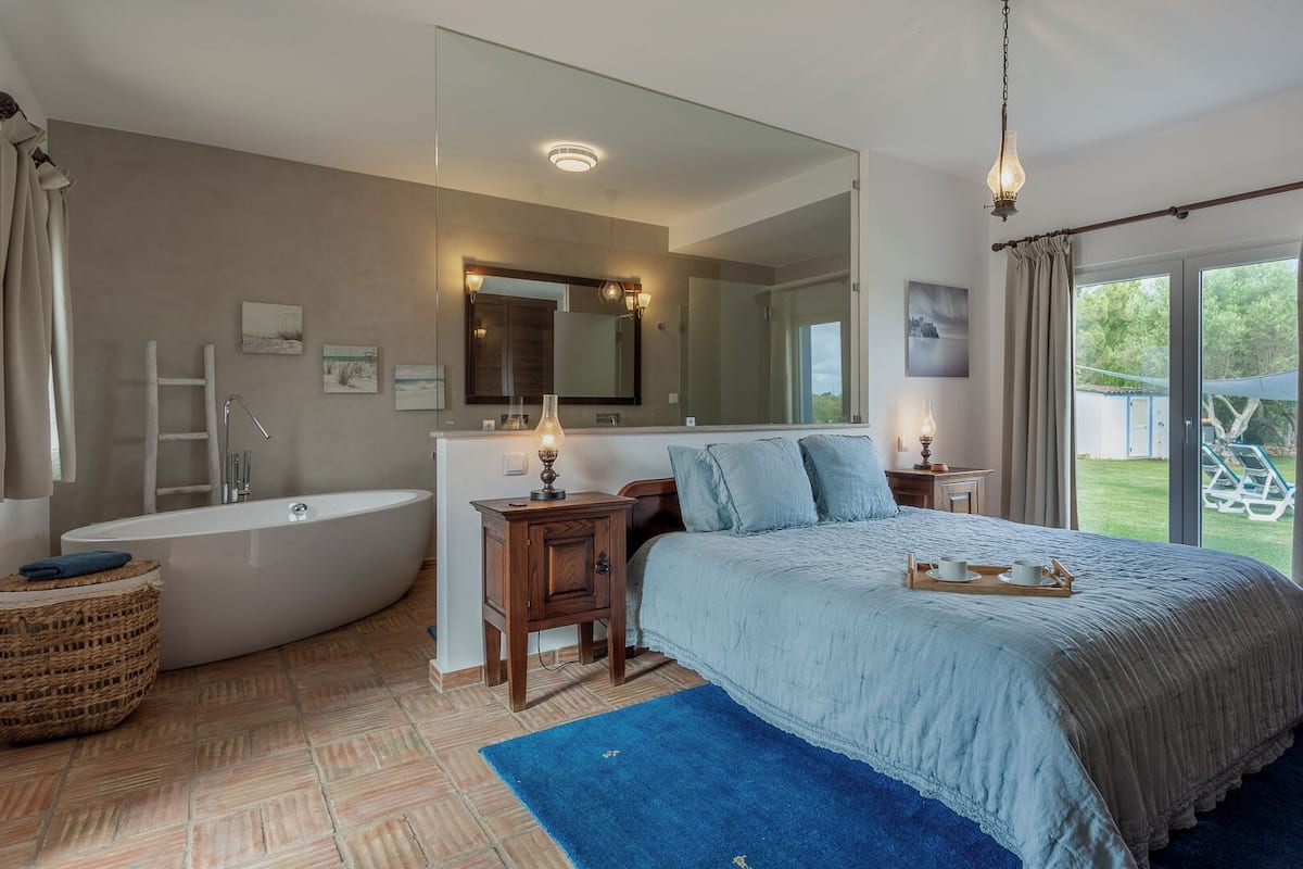 A bedroom with a large bed, a bathtub, a wooden cabinet, a mirror, and a view of the outdoors.