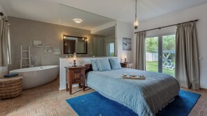 A bedroom with a large bed, a bathtub, a wooden cabinet, a mirror, and a view of the outdoors.