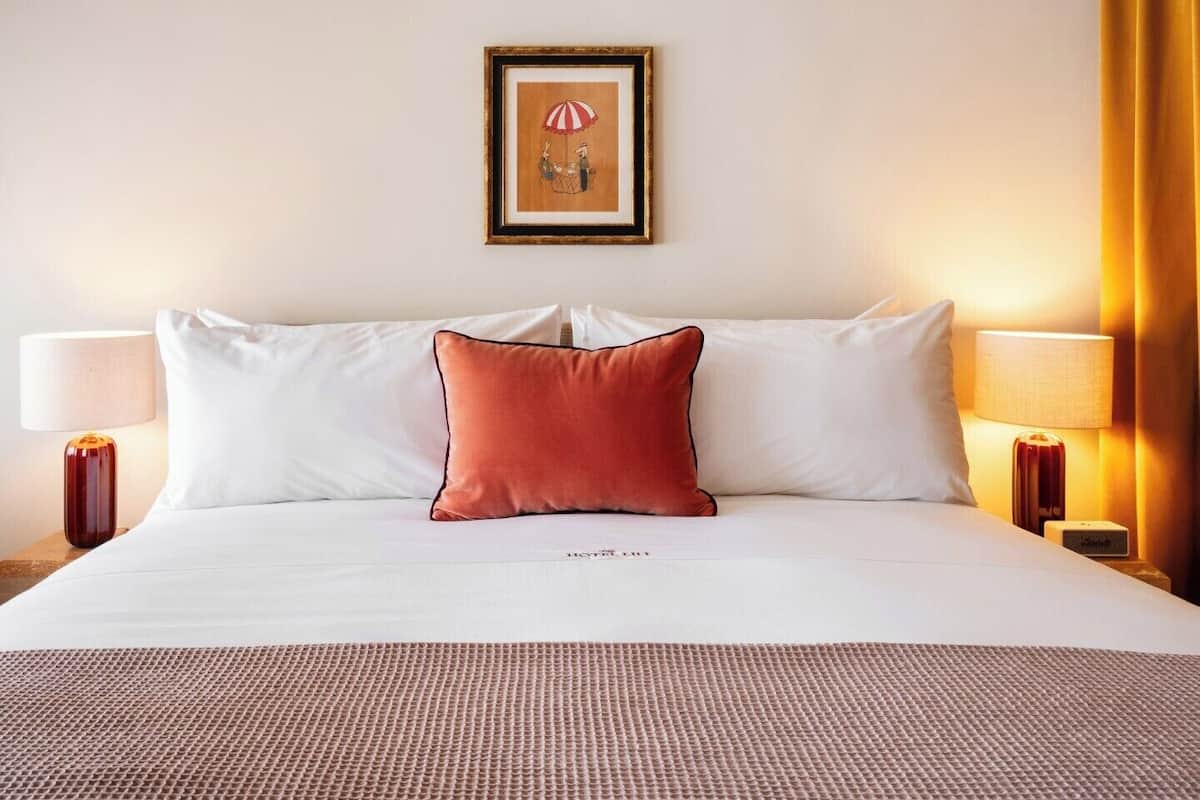 A hotel room with a bed, two bedside lamps, a framed picture on the wall, and a textured rug.