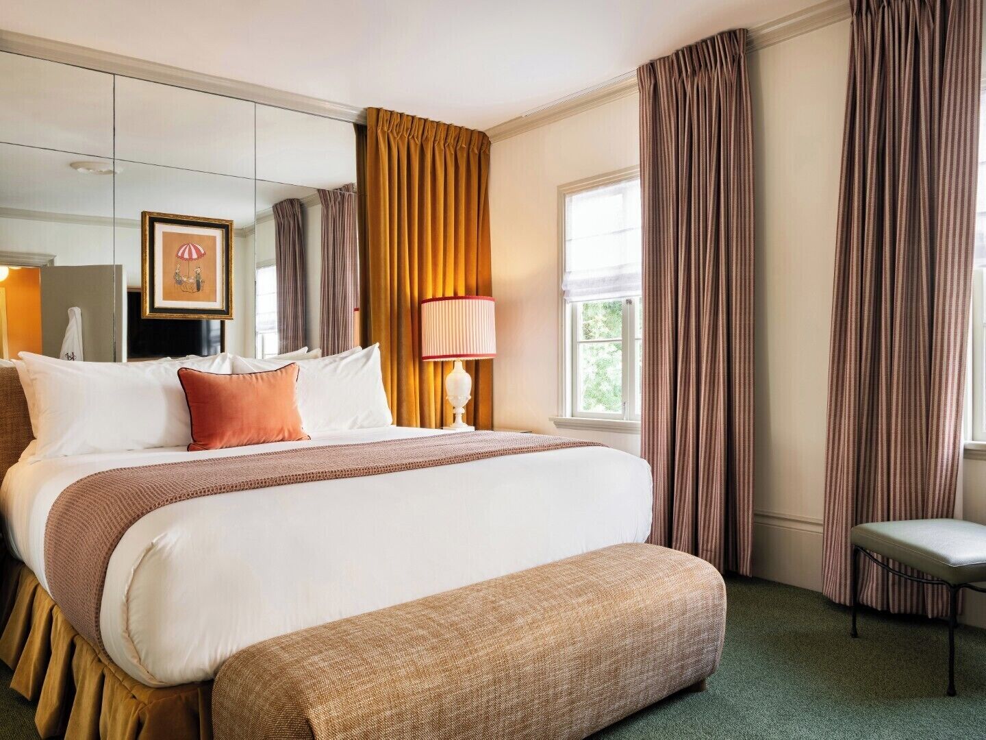 Une chambre d’hôtel avec un grand lit, une table de chevet, une lampe, un banc et une fenêtre avec des rideaux.