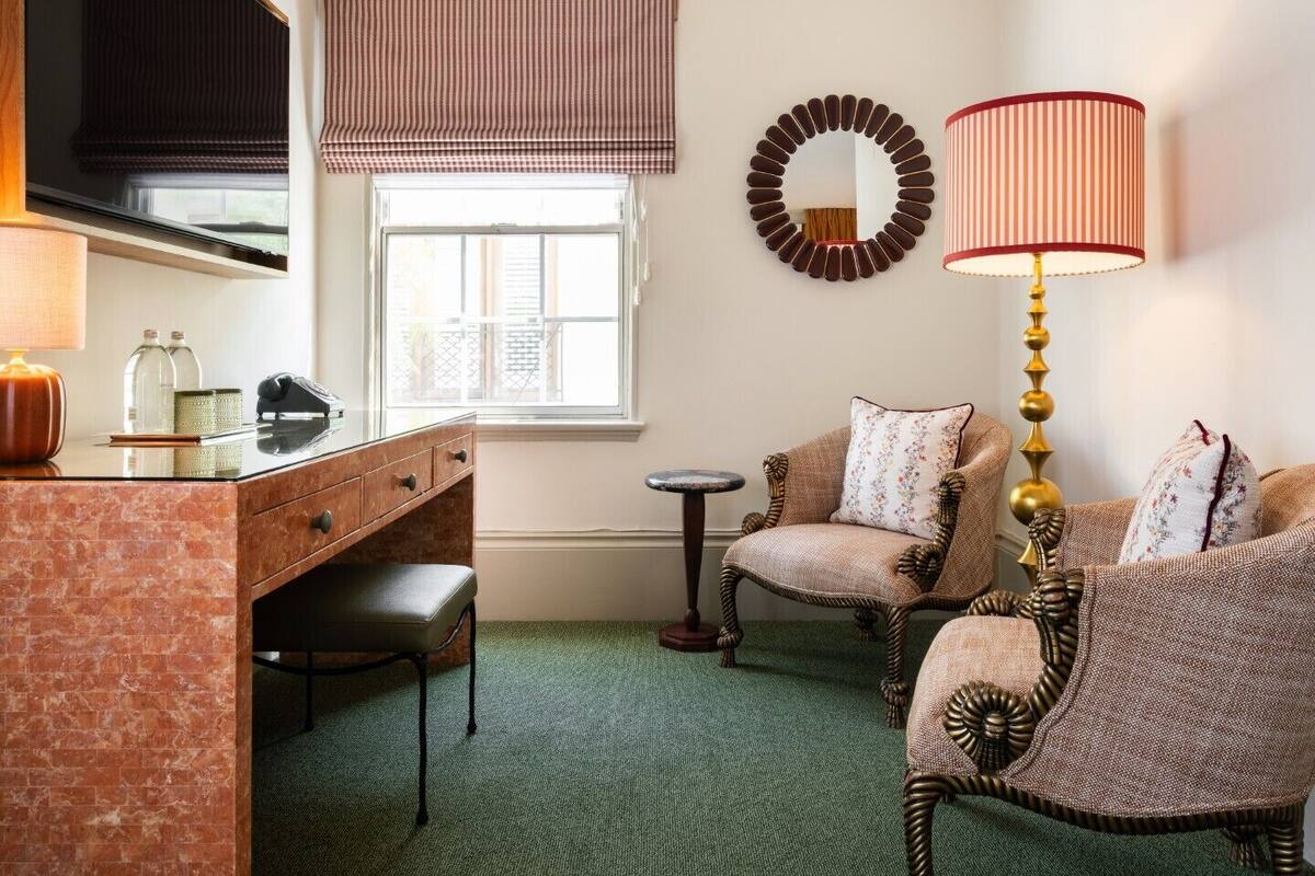 A room with a desk, two chairs, a lamp, a mirror, and a window with blinds.