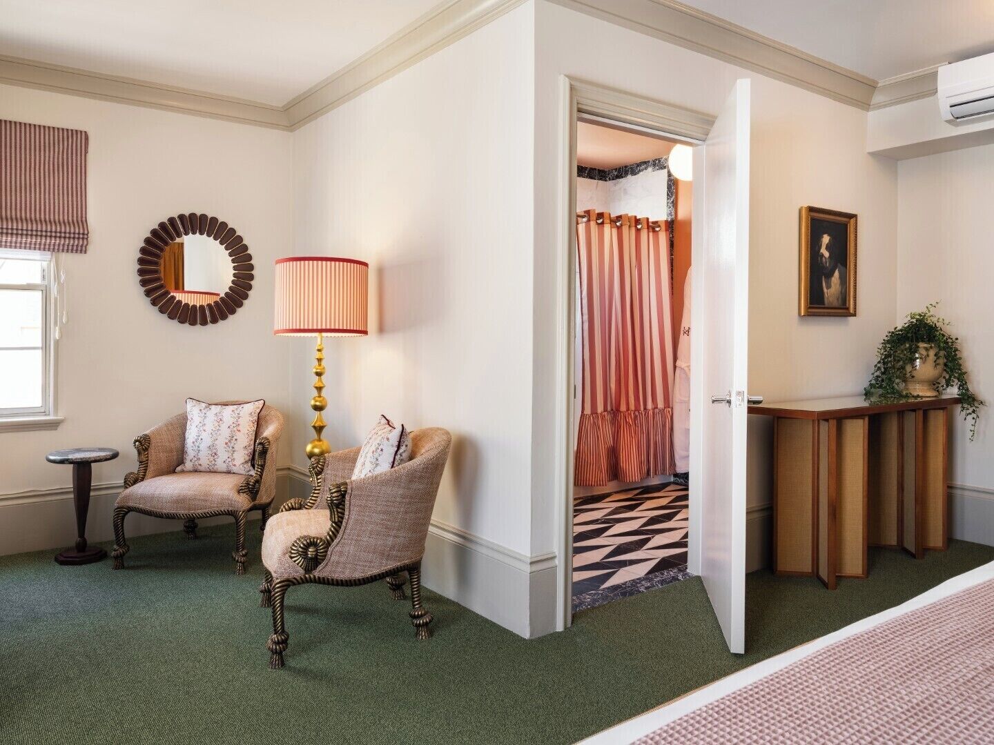 A room with a green carpet, two upholstered chairs, a floor lamp, a mirror, a painting, and a wooden console table with a plant.