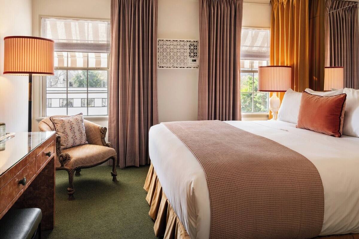 A hotel room with a bed, a desk, a chair, two lamps, and a window with curtains.