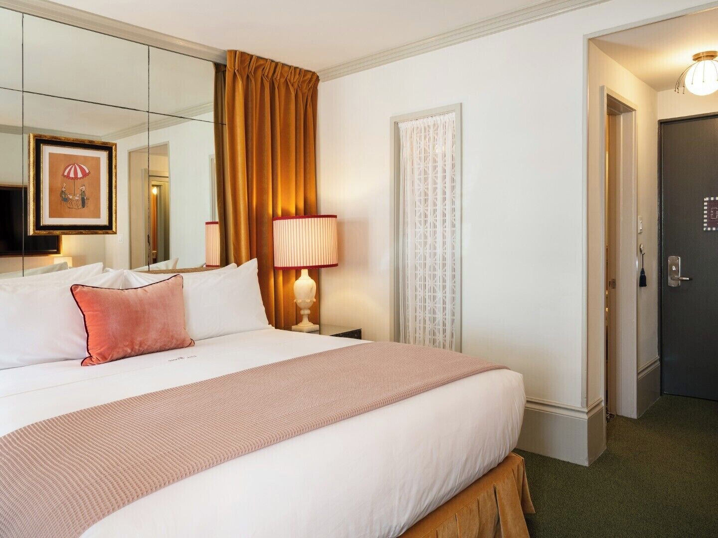 A hotel room with a large bed, a bedside lamp, a mirror, a door, and a framed picture on the wall.