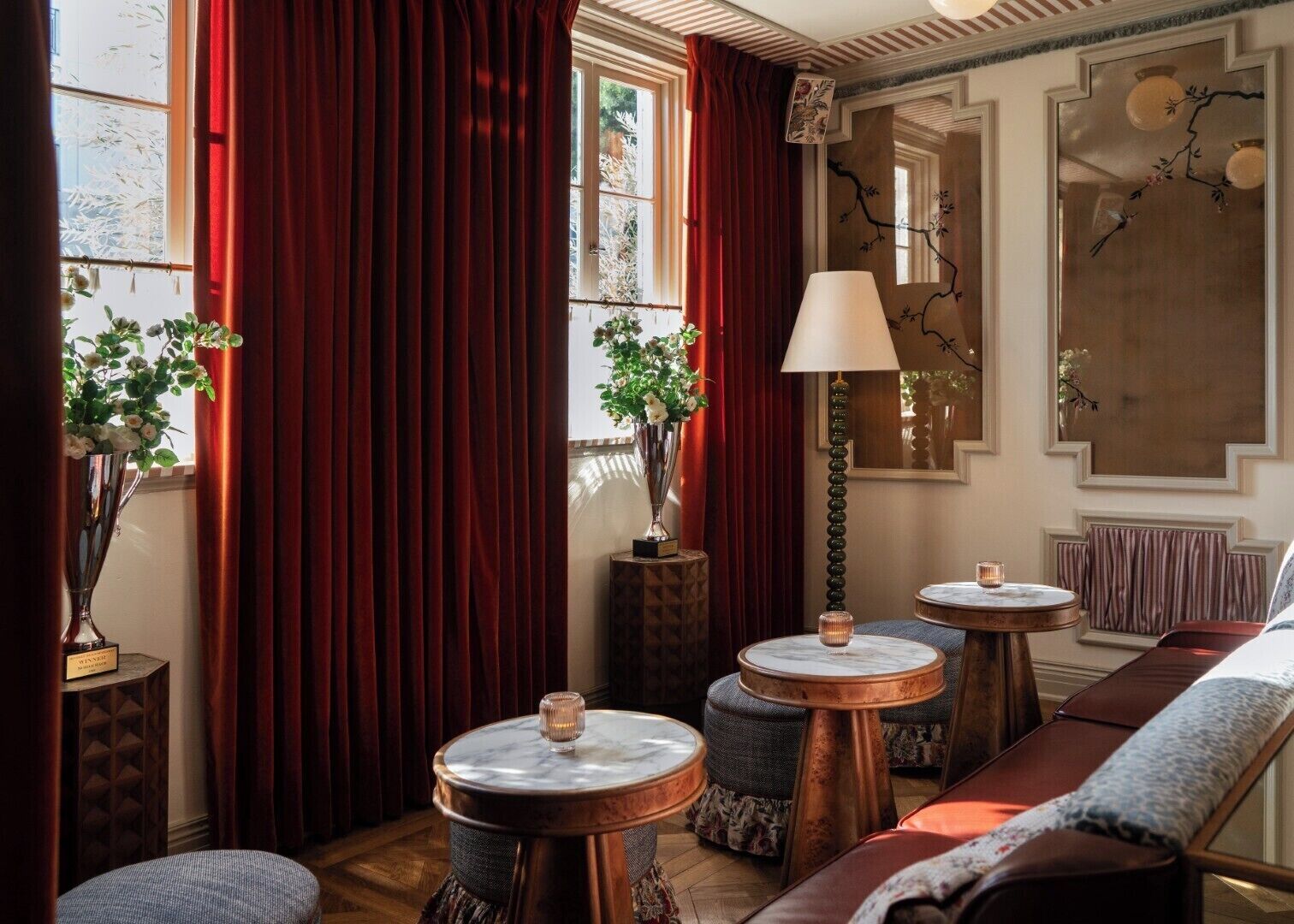 Ein Zimmer mit einer Couch, zwei runden Tischen mit Kerzen, einer Lampe und einem großen Fenster mit Vorhängen.