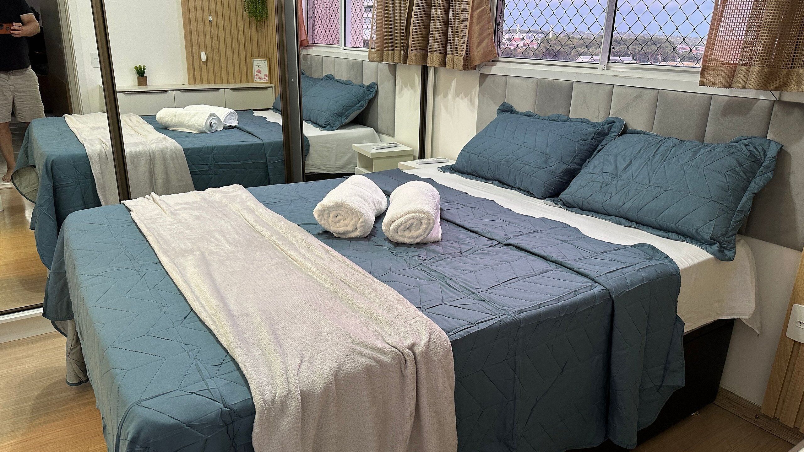 A hotel room with two beds, each with blue bedding and white towels. There is a window with a view of buildings and a plant on the wall.