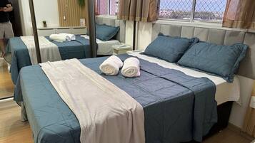 A hotel room with two beds, each with blue bedding and white towels. There is a window with a view of buildings and a plant on the wall.