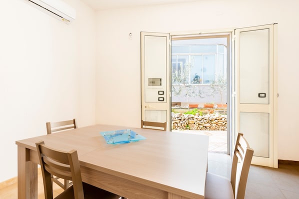 A room with a wooden table, chairs, a sliding glass door to an outdoor area with plants and a stone wall.