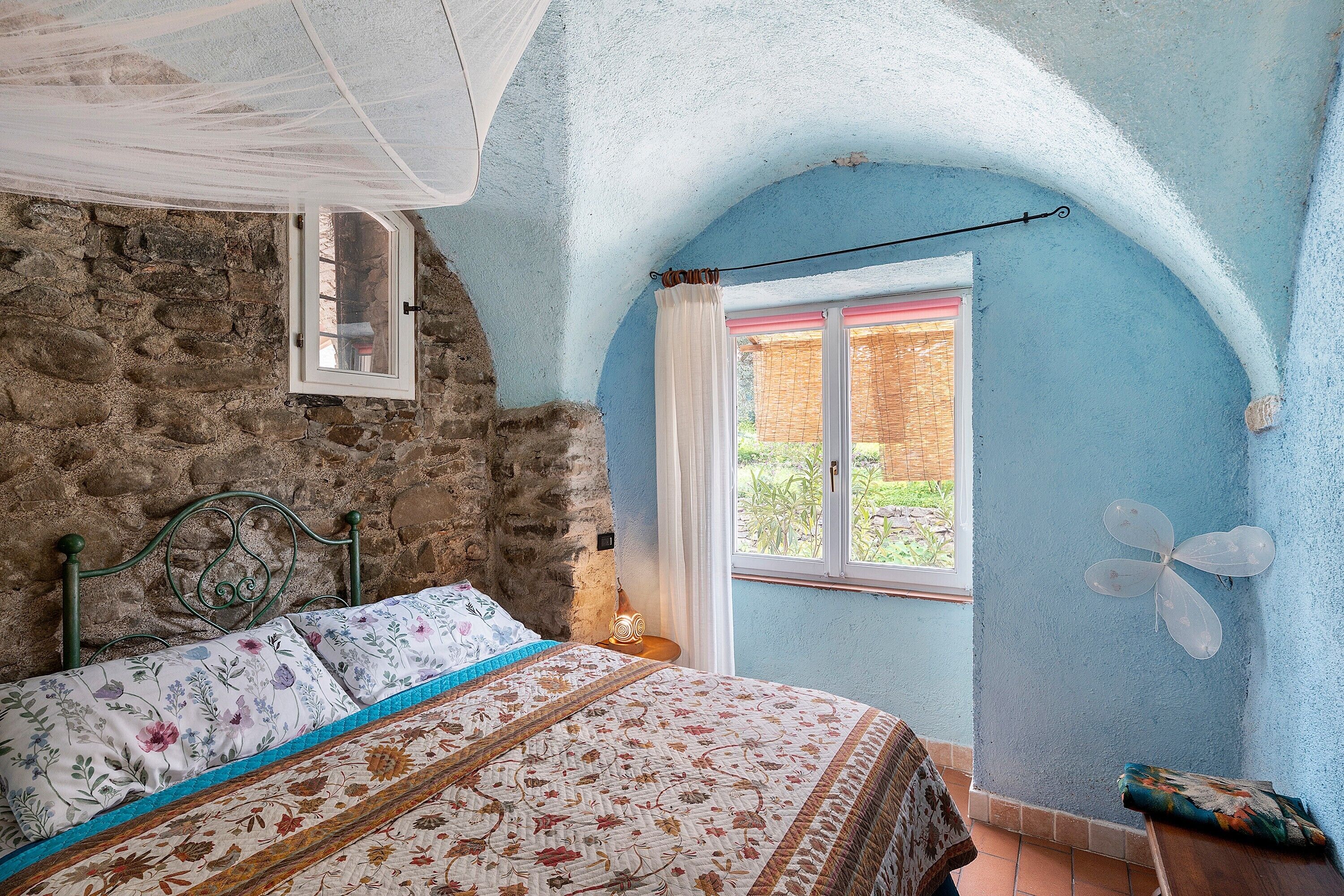 Un dormitorio con pared de piedra, una cama con ropa de cama floral, una ventana con cortina blanca y un ventilador de techo.