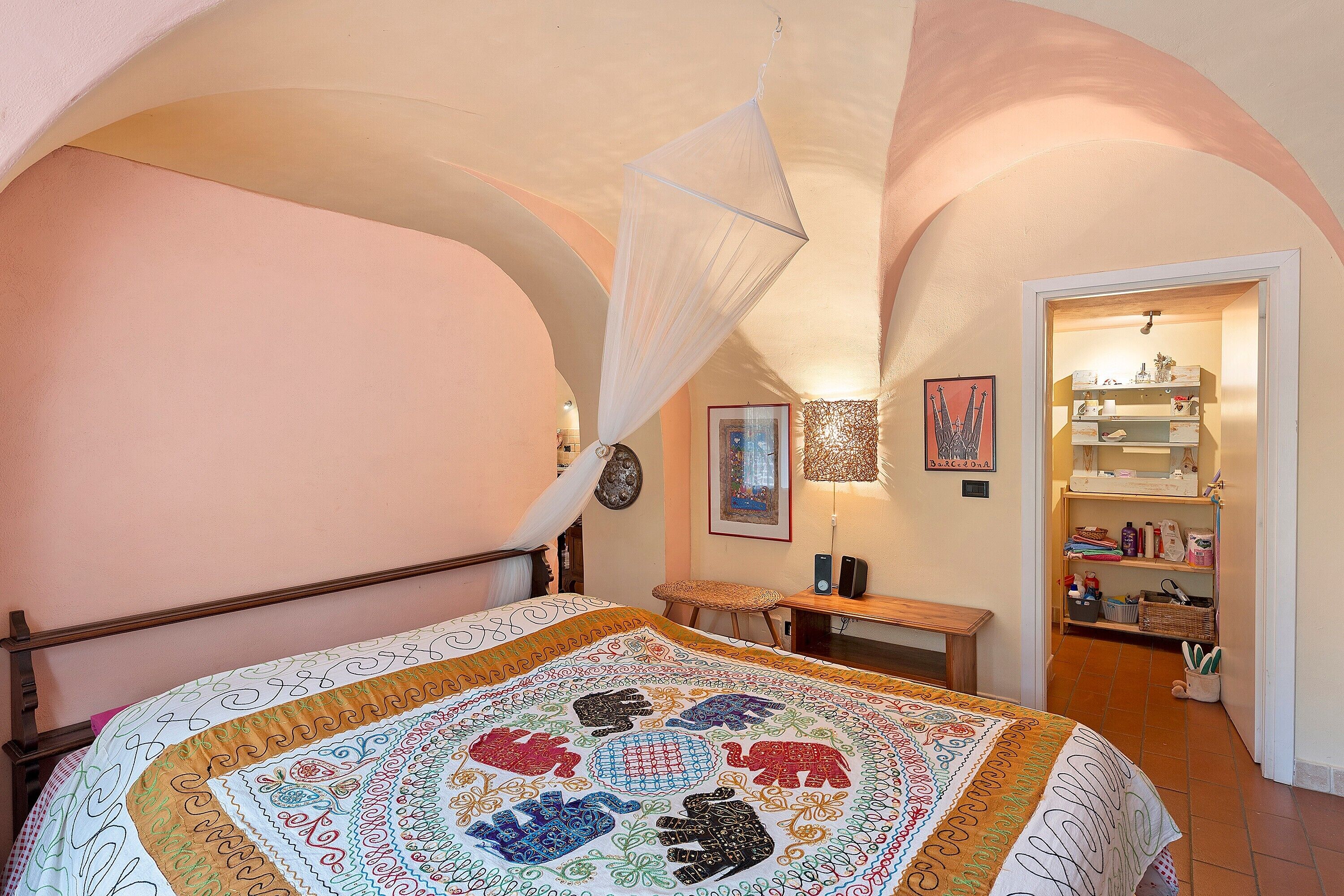 Ein Schlafzimmer mit Bett, einer Bank, einem Nachttisch, einem hängenden Baldachin, einer an der Wand befestigten Uhr, gerahmten Bildern und einer Regalwand mit verschiedenen Gegenständen.