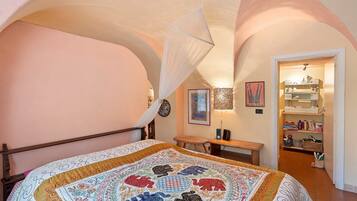 Ein Schlafzimmer mit Bett, einer Bank, einem Nachttisch, einem hängenden Baldachin, einer an der Wand befestigten Uhr, gerahmten Bildern und einer Regalwand mit verschiedenen Gegenständen.