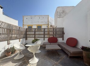 Una terraza en la azotea con un muro blanco, cerca con vallas de madera, y mobiliario, incluyendo una mesa y sillas.