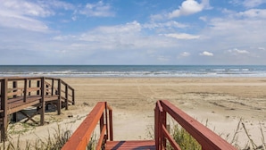 Ein hölzerner Steg führt zu einem Sandstrand mit dem Meer im Hintergrund.
