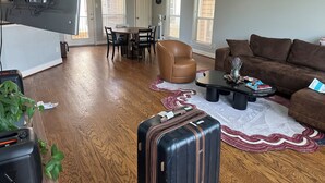 A living room with a brown leather armchair, a brown couch, a dining table with chairs, and a kitchen area with a TV and power outlet. - Cozy 1-bedroom house with AC in quaint Houston neighborhood (Houston)