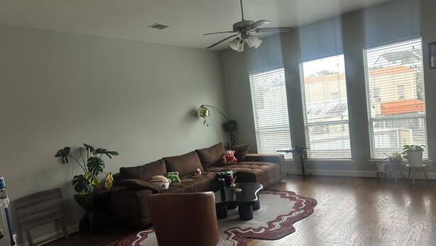 A spacious living room with a brown sofa, a coffee table, and a ceiling fan. There are large windows with blinds, a potted plant, and a small table with a lamp. - Cozy 1-bedroom house with AC in quaint Houston neighborhood (Houston)