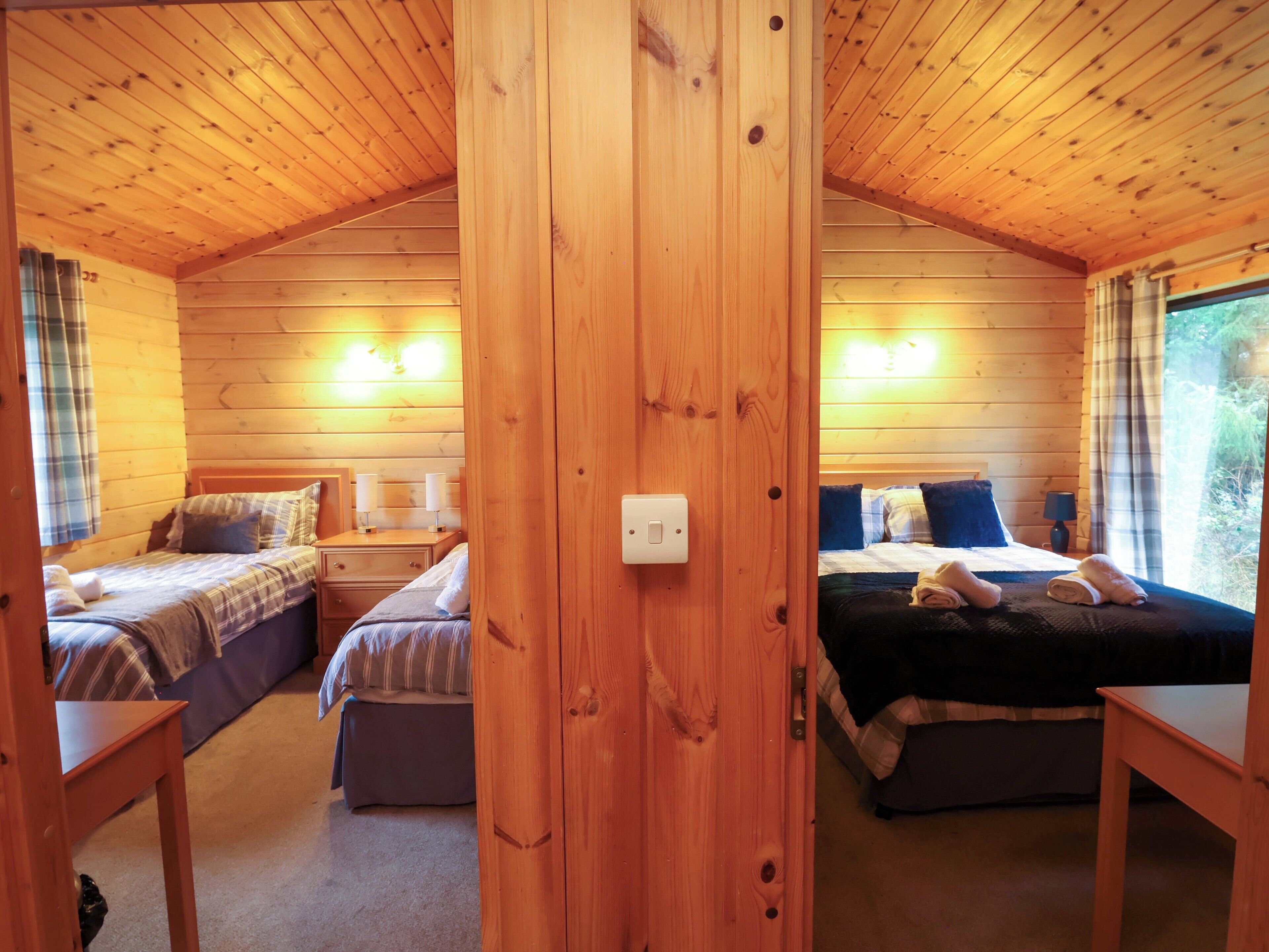 Een chalet met twee slaapkamers, elk met een bed, nachtkastje en lamp. Er is een houten plafond en wanden, en een raam met gordijnen.