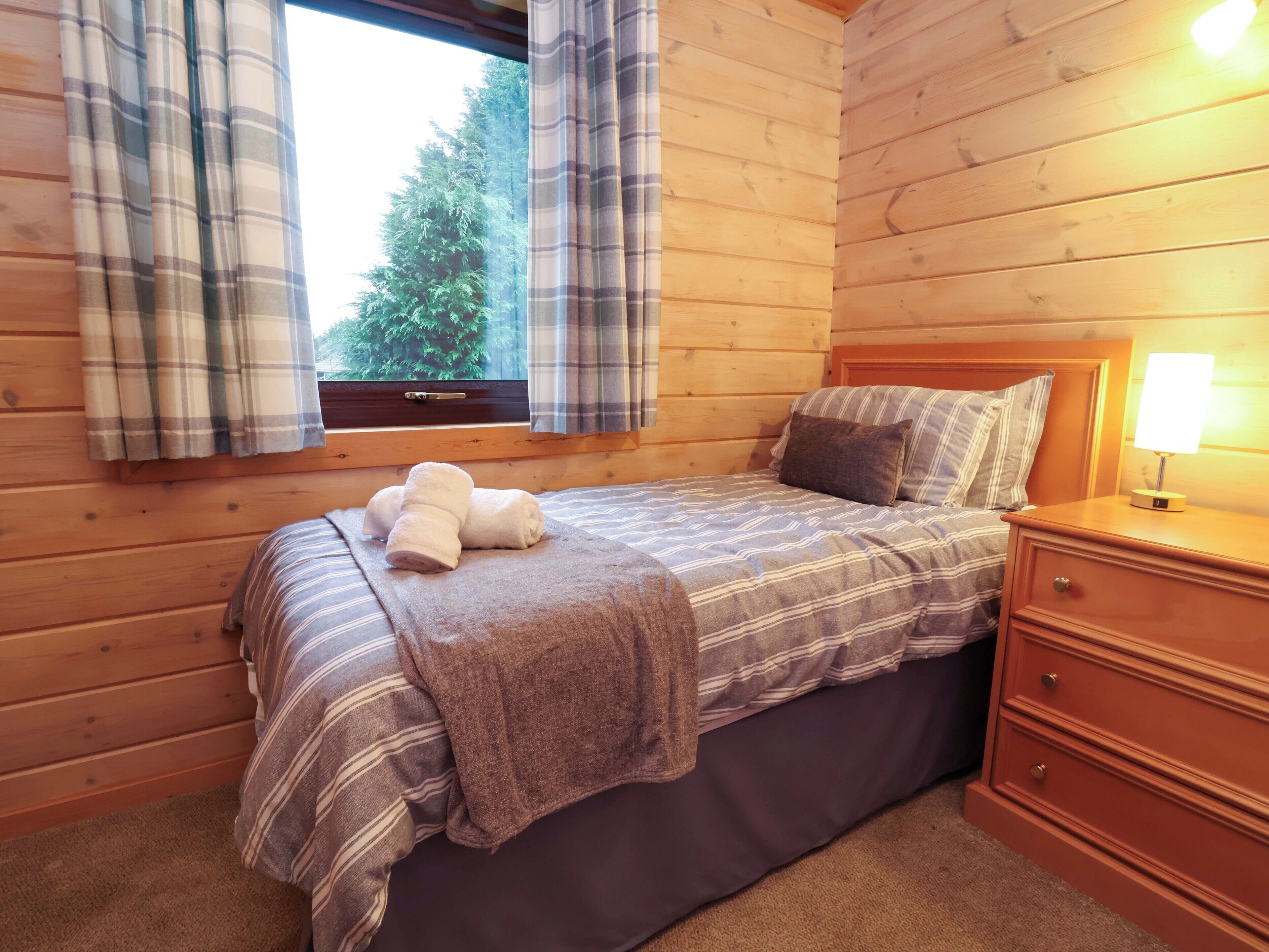 Une chambre dans une cabane en bois, avec un lit, une table de chevet, une lampe et une fenêtre avec des rideaux.