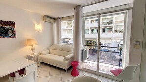 A modern hotel room with a white sofa, a small table with a lamp, a red stool, and a large window offering a view of the city. - HENRI CAMILLE AGENCY- Croisette - 2 bedrooms, 2 Bathrooms, 50m Croisette (Cannes)