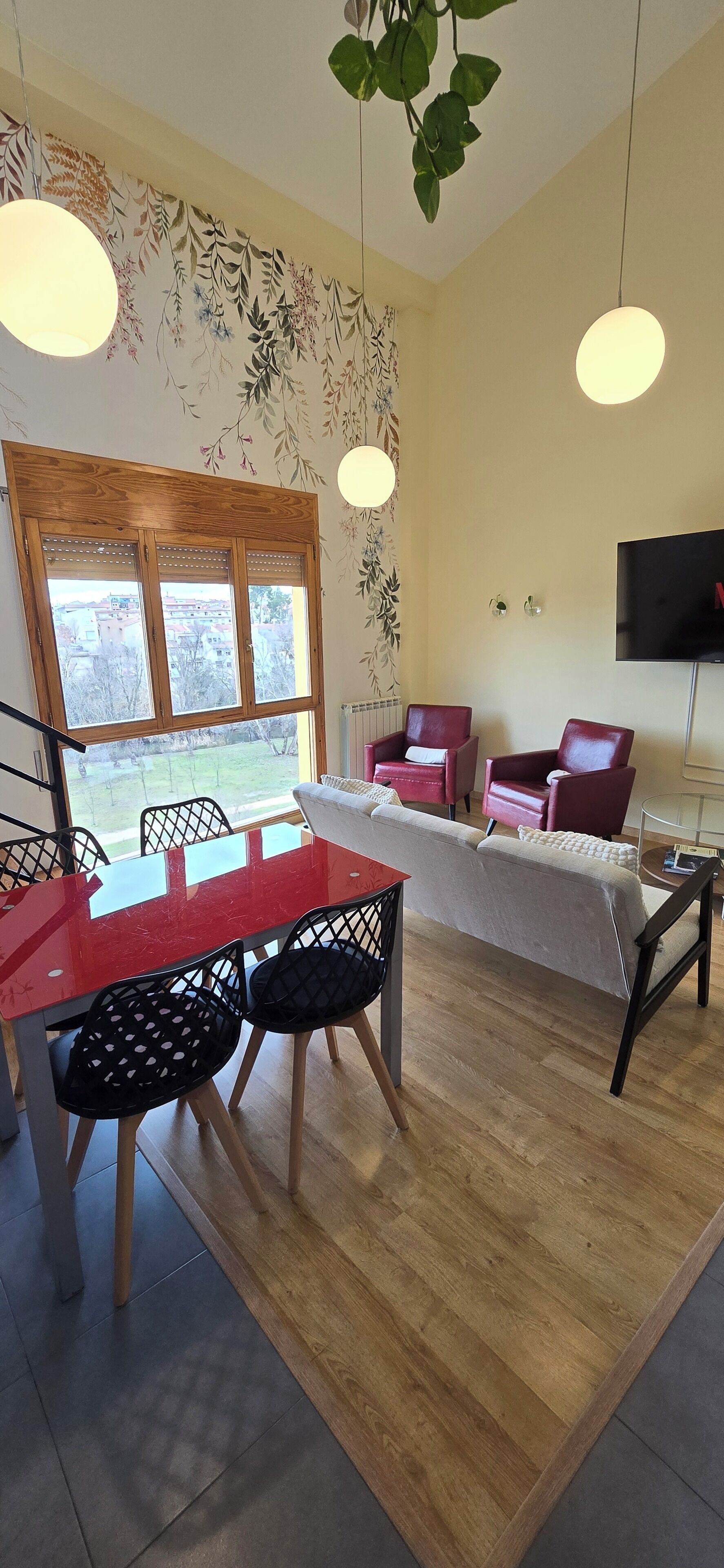 A dining area with a red table and black chairs, a sofa with red cushions, and a TV mounted on the wall.